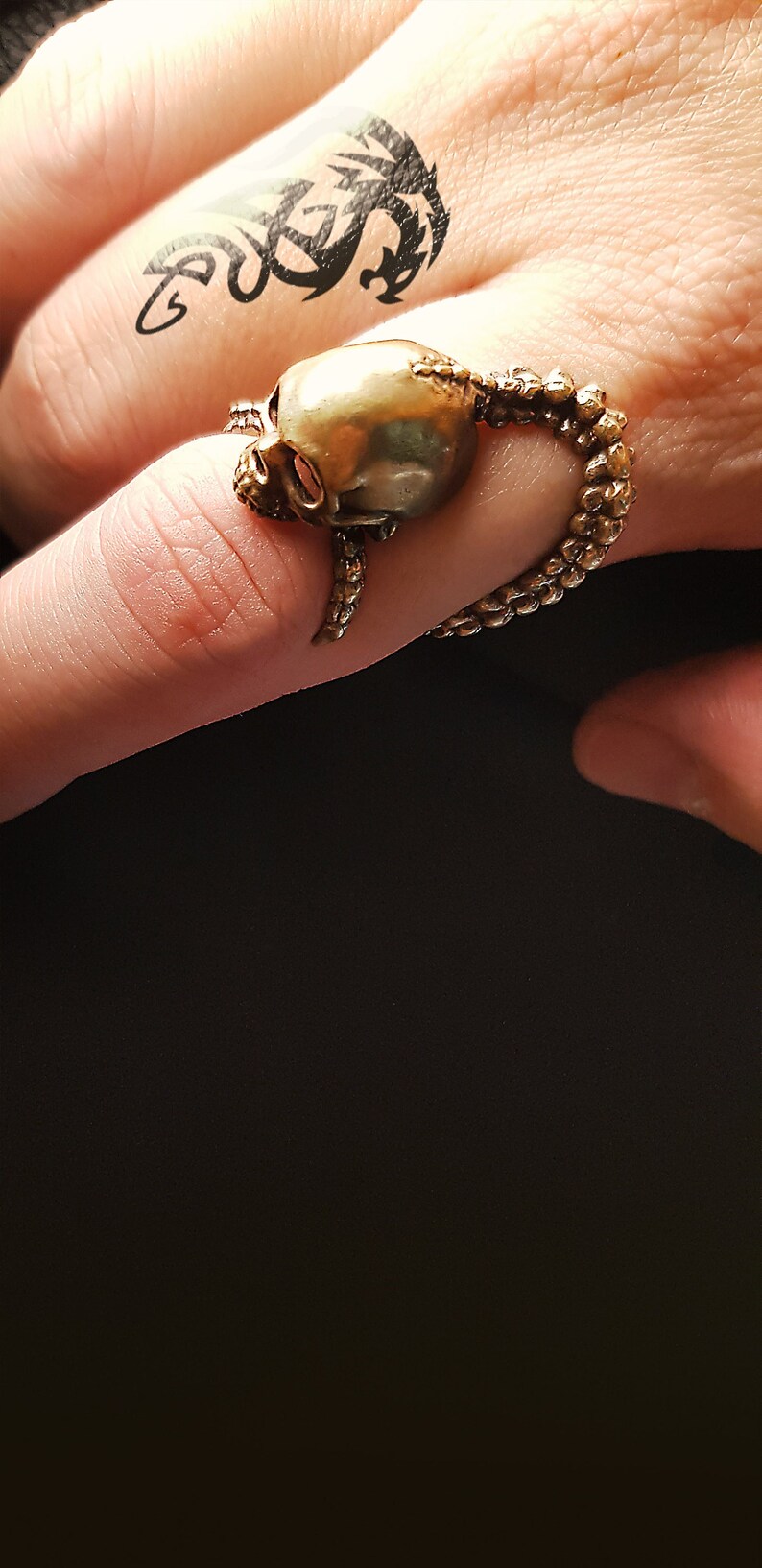 May include: A gold skull ring with a detailed design of tentacles wrapping around the skull. The ring is worn on a finger with a black dragon tattoo.