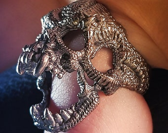 May include: A silver skull ring with intricate details. The ring is worn on a finger and features a detailed skull design with open jaw.