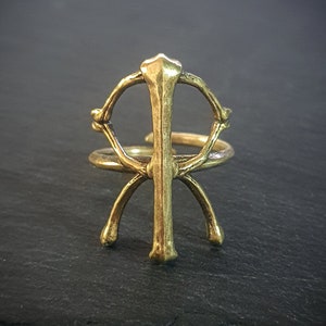 Könnte beinhalten: Ein goldfarbener Ring mit einem einzigartigen Design, das einen Kreis mit einer vertikalen Linie und zwei geschwungenen Linien umfasst, die sich vom Boden erstrecken. Der Ring ist aus Metall und hat eine polierte Oberfläche.