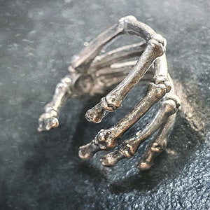 Può includere: Un anello a forma di mano scheletrica in argento con un design dettagliato. L'anello è in metallo e ha un aspetto realistico.