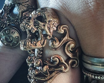 Könnte beinhalten: Ein goldfarbener Ring mit einem detaillierten Totenkopfdesign und filigranen Schnörkeln. Der Ring wird an einem Finger mit anderen Ringen getragen.