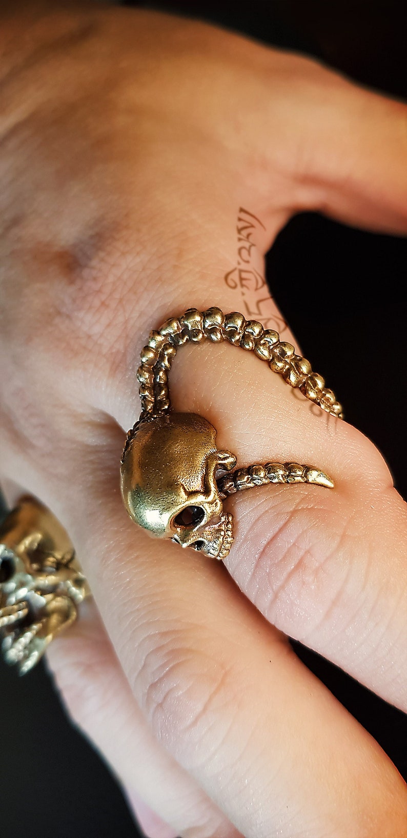 May include: A gold skull ring with a detailed design. The ring is worn on a finger and features a skull with a long, curved horn.