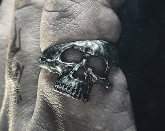May include: A silver skull ring with a detailed design. The ring is worn on a finger.