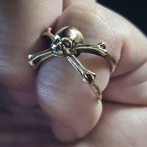 Puede incluir: Un anillo de tono dorado con un diseño de calavera y huesos cruzados. La calavera está detallada con cuencas de los ojos y una nariz.