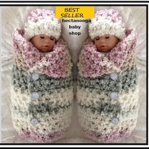May include: Two baby dolls wrapped in a crocheted blanket with a pink, white, and gray color scheme. The blanket has a button detail and the dolls are wearing matching crocheted hats.
