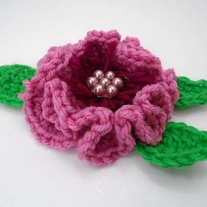 May include: A pink crocheted flower with green crocheted leaves and a center of pearl-like beads.
