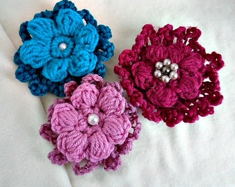 May include: Three handmade crochet flower brooches in shades of teal, fuchsia, and lavender. Each flower features a pearl bead center. The brooches are displayed on a white surface, showcasing the intricate details of the yarn work.