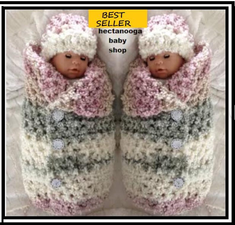 May include: Two baby dolls wrapped in a crocheted blanket with a pink, white and grey colour scheme. The blanket has a button detail and the dolls are wearing matching crocheted hats.