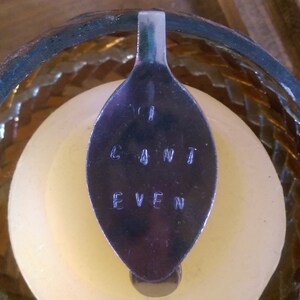 Puede incluir: Una cuchara de plata con las palabras "I can't even" grabadas en ella. La cuchara está apoyada en un portavelas de vidrio transparente.