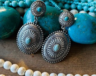 Puede incluir: Un par de pendientes de plata con piedras turquesa. Los pendientes tienen un diseño ovalado grande con una piedra redonda más pequeña en la parte superior. Los pendientes están rodeados de cuentas blancas y turquesa.