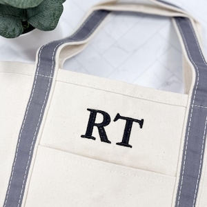 May include: Cream-colored tote bag with gray trim and handles. The bag features a front pocket and the embroidered initials "RT" in black. The handles are reinforced with gray stitching, and the bag appears to be made of canvas.