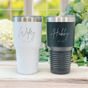 May include: Two insulated tumblers, one white and one black, with silver lids. The white tumbler says "Wifey EST. 2024" in cursive, and the black tumbler says "Hubby EST. 2024". Both tumblers have a ridged design.