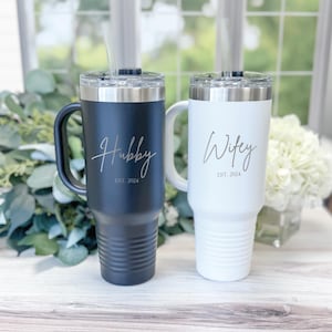 May include: Two stainless steel travel mugs with handles. The black mug says "Hubby Est. 2024" and the white mug says "Wifey Est. 2024".