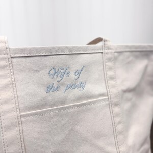 K&ouml;nnte beinhalten: Eine wei&szlig;e Canvas-Tragetasche mit einem blauen gestickten Spruch "Wife of the party" auf der vorderen Tasche.