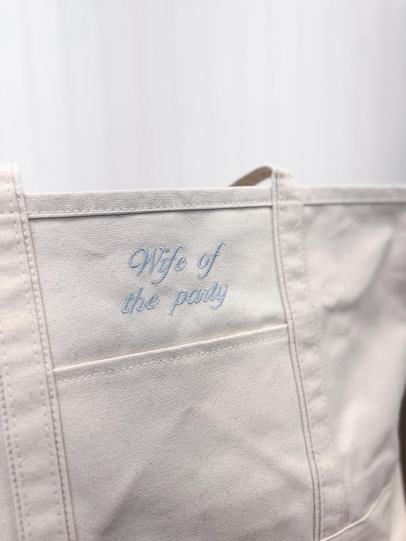 K&ouml;nnte beinhalten: Eine wei&szlig;e Canvas-Tragetasche mit einem blauen gestickten Spruch "Wife of the party" auf der vorderen Tasche.