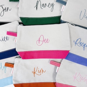 May include: Nine personalized makeup bags with different names embroidered on them. Each bag is made of white canvas with a colored bottom. The names are Dee, Jen, Nancy, Rice, Kim, Krist, Ragan, and Vii.