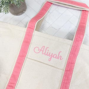 May include: Cream-colored tote bag with pink handles and trim. The name "Aliyah" is embroidered in pink script on the front pocket. The bag has a simple, classic design, suitable for everyday use.
