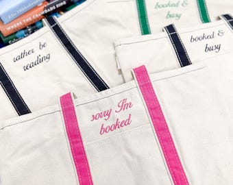 May include: Three canvas tote bags with different coloured handles and embroidered text. The top bag has green handles and says "booked & busy". The middle bag has black handles and says "booked & busy". The bottom bag has pink handles and says "sorry I'm booked".