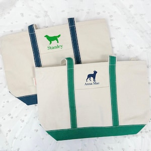 May include: Two canvas tote bags with green and navy blue trim. The larger bag has a green silhouette of a dog with the name "Stanley" embroidered on it. The smaller bag has a navy blue silhouette of a dog with the name "Anna Mae" embroidered on it.
