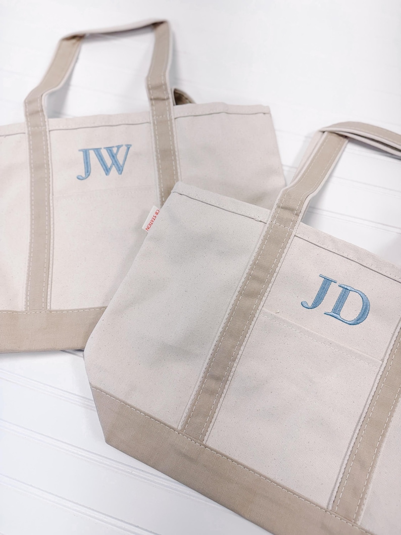 K&ouml;nnte beinhalten: Zwei beigefarbene Canvas-Tragetaschen mit braunem Besatz. Die Tasche links hat die Initialen "JW" in Blau gestickt. Die Tasche rechts hat die Initialen "JD" in Blau gestickt.