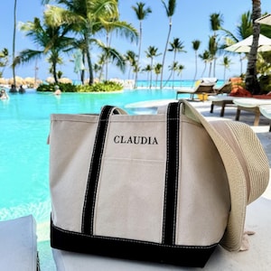 May include: A white canvas tote bag with black straps and a black bottom. The bag has the name "CLAUDIA" embroidered on the front in black.