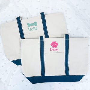 May include: Two white canvas tote bags with navy blue handles and trim. The top bag has a green bone and the name "Bella" embroidered on it. The bottom bag has a pink paw print and the name "Daisy" embroidered on it.