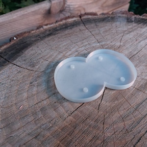 May include: A light blue, two-part resin coaster with four small, raised circular feet. The coaster is shaped like two connected circles and sits on a weathered, textured wooden surface.