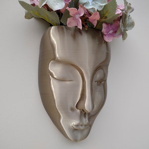 Peut inclure: Un pot mural en forme de visage, de couleur dorée, avec les yeux fermés et la bouche légèrement entrouverte. Le pot est rempli de fleurs roses et vertes.