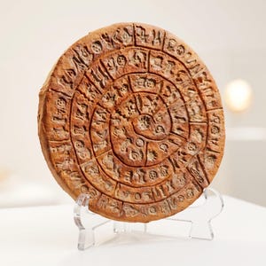 Peut inclure: Un disque en argile de couleur terre cuite, de forme circulaire, présentant un motif en spirale composé de symboles pictographiques estampés sur les deux faces. L'objet est posé verticalement sur un support en acrylique transparent.