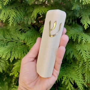 Op de afbeelding: Een beige mezuzah-koker met een goudkleurige Hebreeuwse letter Shin. De rechthoekige koker wordt tegen een achtergrond van groen gebladerte gehouden. De mezuzah-koker is een religieus item.