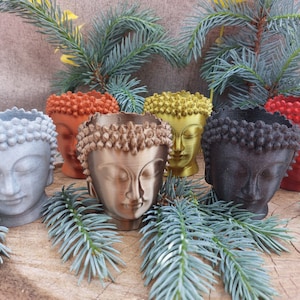 Puede incluir: Seis pequeños maceteros con forma de cabeza de Buda en diferentes colores, incluyendo gris, marrón, dorado, rojo y negro. Los maceteros están hechos de un material liso y texturizado y están dispuestos sobre una superficie de madera.