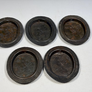 May include: A set of five circular dark brown metal coasters with a ribbed edge and a recessed center featuring an embossed landscape scene with figures and trees. Each coaster has a uniform dark patina and a distinct textured border design.