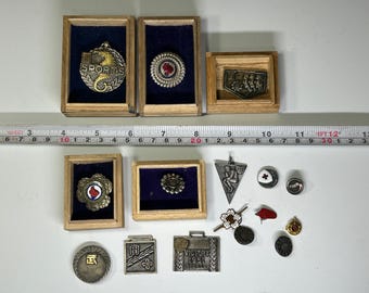 Op de afbeelding: Een verzameling vintage metalen pinnen en badges in verschillende ontwerpen, waaronder een sportbadge met het woord "SPORTS" in het midden, een badge met een rood kruis en een badge met het woord "VICTORY" en het jaar "1951".