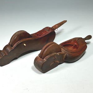 Puede incluir: Dos antiguas herramientas de marcado de madera, probablemente para carpintería o ebanistería. Las herramientas son de color marrón oscuro con un interior rojizo, con una bobina y un mango. Una herramienta tiene una cuerda que se extiende.