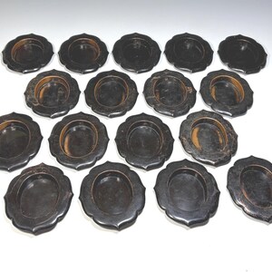 May include: A collection of fourteen small, oval-shaped black lacquer coasters or dishes. Each piece features a scalloped edge design with a smooth, dark finish and a slightly recessed center, arranged in a grid pattern on a plain surface.