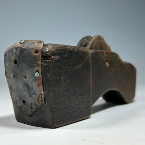 Puede incluir: Zueco de madera oscura con una placa metálica en el talón asegurada con remaches. La placa metálica muestra signos de desgaste. El zueco tiene una suela curva y un borde superior decorativo.