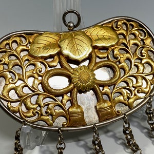 May include: A silver and gold ornate pendant with a floral design and a bow. The pendant has a loop at the top for hanging and five chains hanging from the bottom.