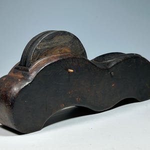 Puede incluir: Una herramienta de madera marrón oscuro con forma redondeada y dos discos circulares negros en la parte superior. La herramienta tiene un pequeño mango de madera que se extiende desde el lateral.