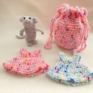 May include: A collection of handmade crochet items, including a gray mouse, a pink drawstring bag, and two small dresses, one pink and one blue. The items are decorated with colorful flecks.
