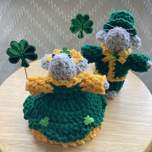 May include: Two crocheted mice dressed in green and yellow outfits with shamrock accents. The mice are holding shamrock decorations.