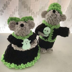 May include: Two crocheted mouse figures, one in a black dress with a ghost button, the other in a black jacket with a skull button. Both mice have gray bodies, green accents, and green hats, perfect for Halloween.