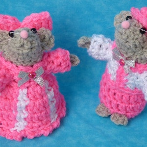 May include: Two handmade crochet mice, one with a large pink bow and the other with a pink hat. Both mice wear pink and white outfits with silver bows. The mice have gray bodies, pink noses, and black eyes. The background is a solid light blue.