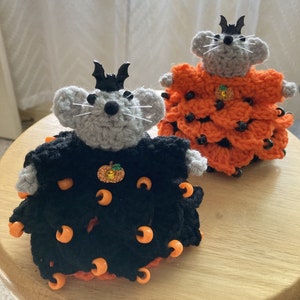 May include: Two crocheted mice dressed in Halloween costumes. One mouse is wearing a black dress with orange beads and a pumpkin charm. The other mouse is wearing an orange dress with black beads.