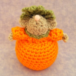 May include: A crocheted stuffed animal shaped like a mouse wearing a bright orange pumpkin costume. The mouse has light tan yarn fur, a small pink nose, and long thin whiskers. It is topped with a textured green crocheted leaf collar.