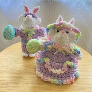 May include: Two crocheted Easter bunny figurines, one in a purple dress with a green and white hat and the other in a purple and white outfit, holding a green and white speckled Easter egg.