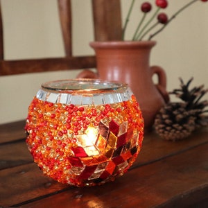 May include: A round glass candle holder with a mosaic design in shades of red, orange, and yellow. The candle holder is made of small glass tiles and beads.