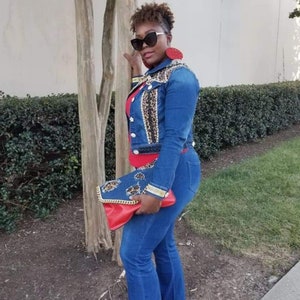 May include: A woman wearing a blue denim jacket with leopard print and gold accents, blue jeans, and red earrings. She is holding a red and blue clutch with a gold chain.