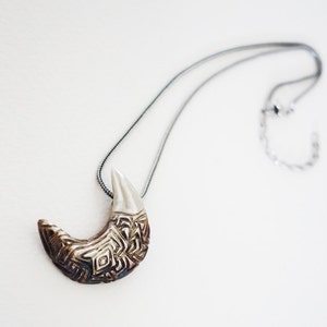 May include: A silver chain necklace with a crescent moon pendant. The pendant is made of metal and has a textured, geometric design in shades of brown and white.