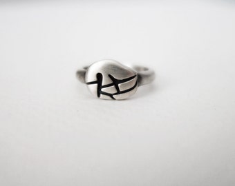 Puede incluir: Un anillo de plata con una parte superior redonda y texturizada que presenta un diseño abstracto negro.