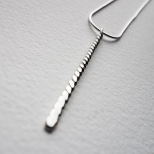 May include: A silver necklace with a long, thin pendant made of small, round beads. The pendant is twisted and hangs straight down.